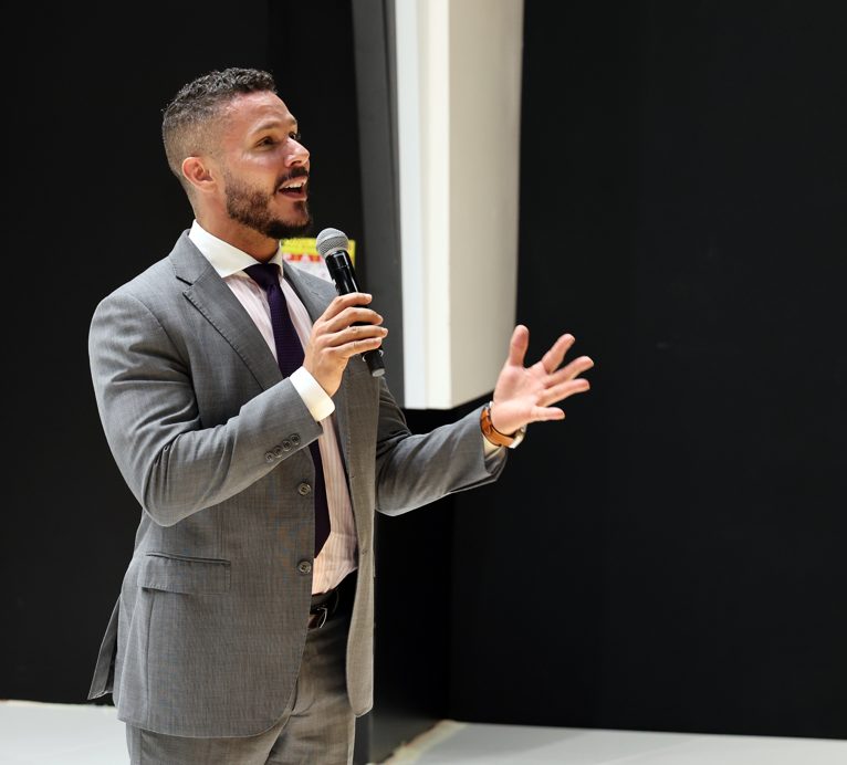 Andrew Wolhuter speaking on stage with a microphone against a dark backdrop